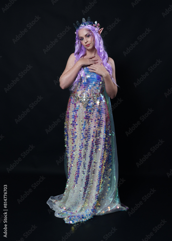 Full length portrait of beautiful female model with long purple hair wearing elf ears, a fantasy fairy crown and  rainbow glitter sequin ball gown. graceful standing pose,  isolated in dark studio bac