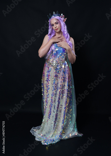 Full length portrait of beautiful female model with long purple hair wearing elf ears, a fantasy fairy crown and rainbow glitter sequin ball gown. graceful standing pose, isolated in dark studio bac