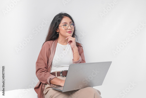 Thoughtful young Asian business woman sitting down with a laptop thinking about work, isolated by white background.