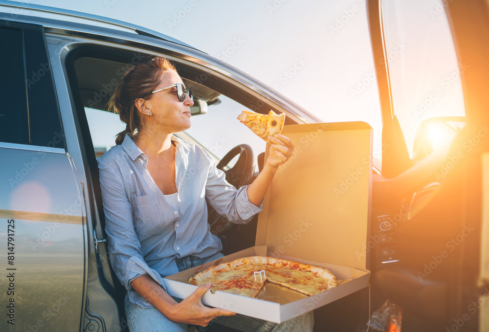 Naklejka premium Midle-aged woman eating just cooked italian pizza sitting on driver car seat during meal break and enjoying sunset light. Auto traveling, fast food eating or car jorney lunch break concept image.