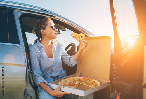 Midle-aged woman eating just cooked italian pizza sitting on driver car seat during meal break and enjoying sunset light. Auto traveling, fast food eating or car jorney lunch break concept image. photo