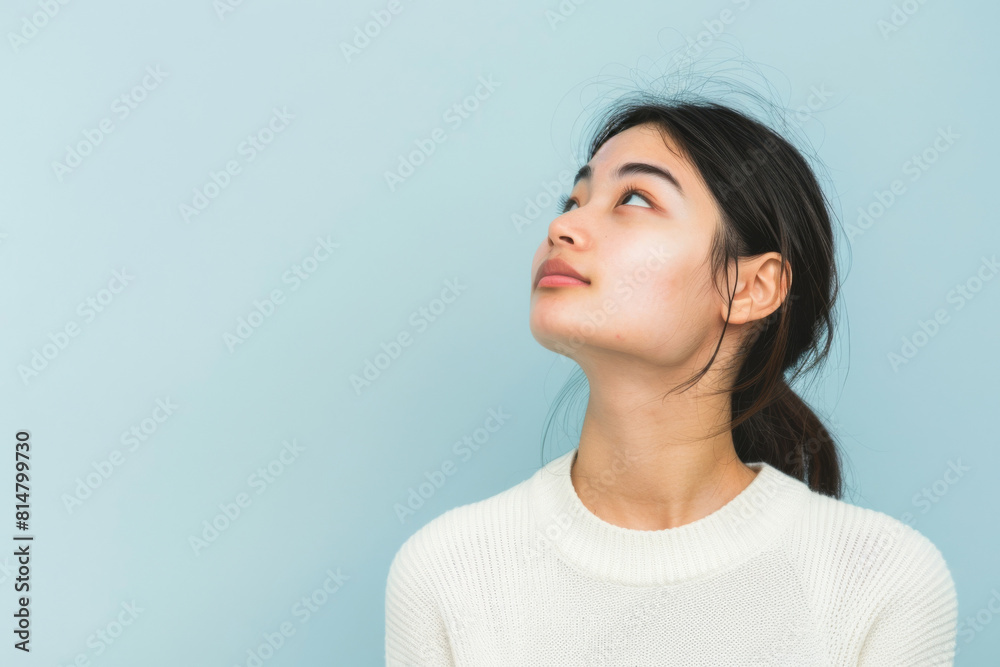 Thoughtful young woman in a white sweater looks upwards while standing against a solid blue studio background, lost in contemplation Thoughtful young woman in a white sweater looks upwards while standing against a solid blue studio background, lost in contemplation