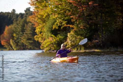 Woman kayaking with autumn foliage on pond or lake with blonde hair and orange boat and purple jacket in Vermont and New England.