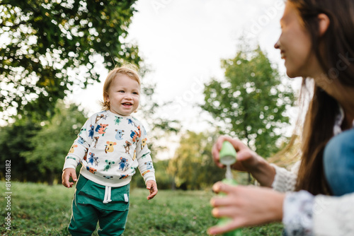 Mom blow soap bubbles outdoor. Toddler little cute walking and playing in spring garden. Boy catches soap bubbles on green grass in park enjoying sunny summer day. Outdoors creative activities for kid