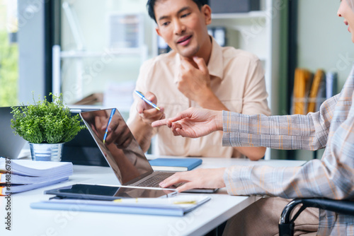 A diverse group of professionals, including a male Asian businessman and a female Muslim businesswoman in hijab, gather around a desk equipped with tablets and laptops for a productive meeting. photo