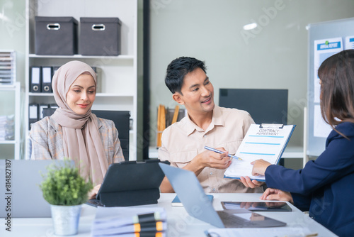 A diverse group of professionals, including a male Asian businessman and a female Muslim businesswoman in hijab, gather around a desk equipped with tablets and laptops for a productive meeting.