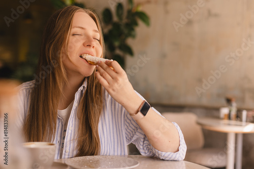Young blonde woman eating eclair sitting in cafe. Girl bite piece of croissant look joyful at restaurant. Cheat meal day concept. Woman is preparing with appetite to eat eclair. Enjoy coconut bakery.