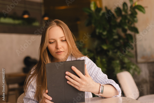 Attractive calm smiling blonde woman wear shirt sitting with tablet computer in cafe. Beauty girl student using tablet computer for social media, studying or reading news.