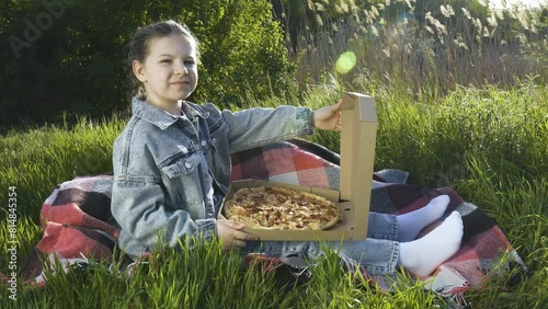 A girl in a blue denim suit sits on a blanket on the green grass and opens a box of pizza. Summer sunny day. Outdoor picnic