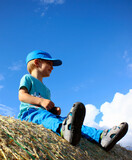 Happy 3-year old Caucasian boy sitting on the top of a hay bale on a summer day with blue sky. Concept for happy carefree childhood, enjoying childhood and summer, Children's Day and wheat harvest end