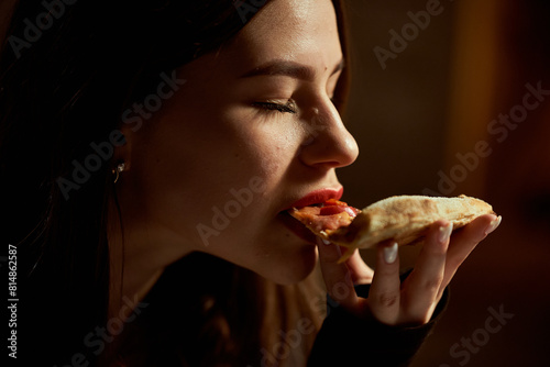 A beautiful girl with her eyes closed enjoys eating pizza. Lips are painted with red lipstick.