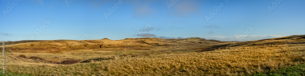Panoramic view of a dry barren grassland prairie with small hills under a bright blue sky.
