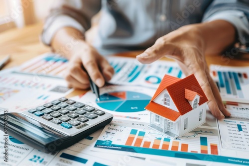 Person holding and pointing to a small house model on a table with a calculator, with documents