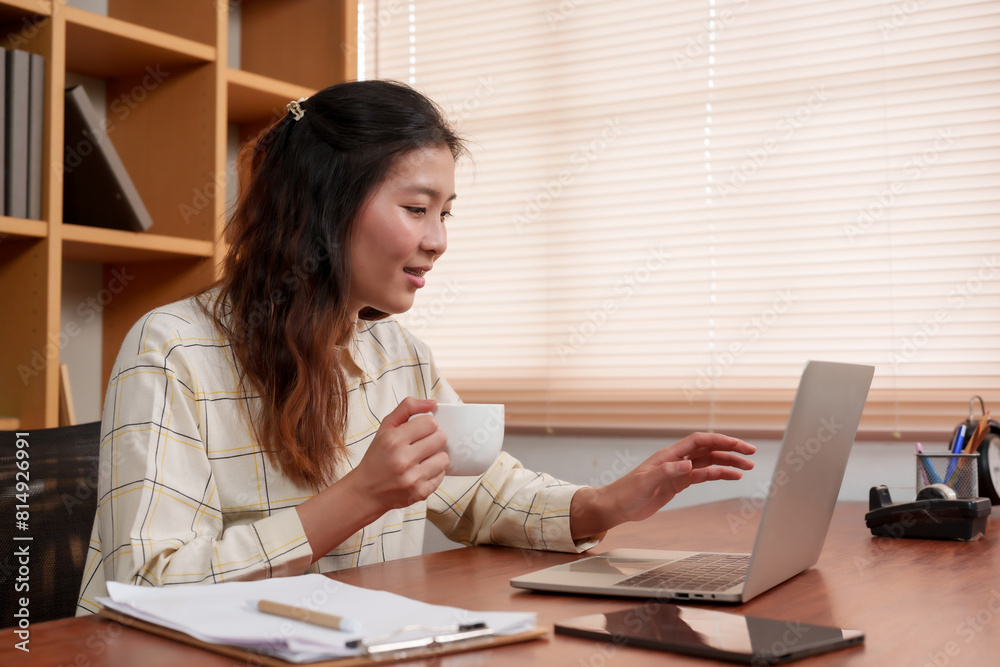 Asian woman holding cup of coffee while working on laptop at desk. Wearing checkered shirt, smiling, in bright office with wooden shelves, documents, and stationery on desk. Asian woman holding cup of coffee while working on laptop at desk. Wearing checkered shirt, smiling, in bright office with wooden shelves, documents, and stationery on desk.