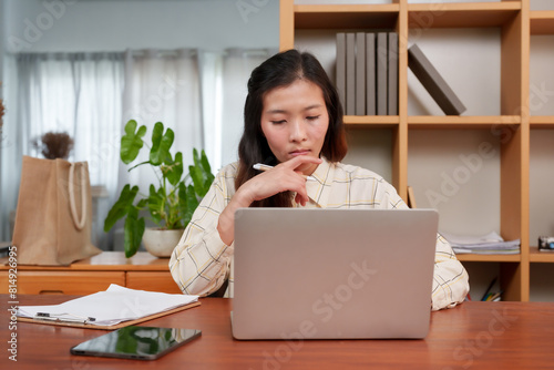 Focused Asian woman in checkered shirt sitting at desk working on laptop in bright office. Holding pen, thinking, surrounded by documents, plants, shelves, and natural light from window.