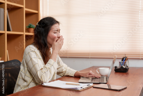 Young Asian woman yawning, covering mouth, feeling tired. Sitting at desk with laptop, documents, coffee cup, bright window blinds in background. Casual checkered shirt, demonstrating work fatigue © Ekkasit A Siam