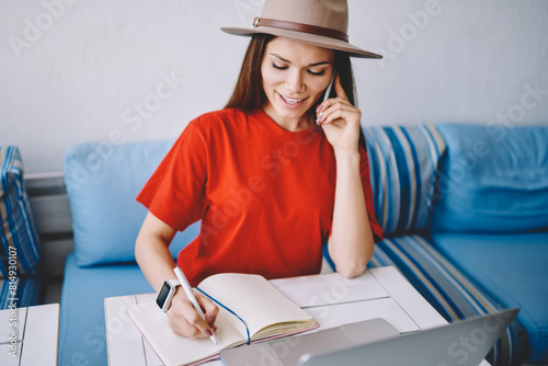 Positive female student calling to customer service for consultancy and writing main theses in textbook, successful woman checking account balance via telephone conversation and notes information photo