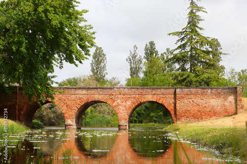 red brick bridge with visible masonry and the reflection of its three arches on the water of a calm river near a forest photo