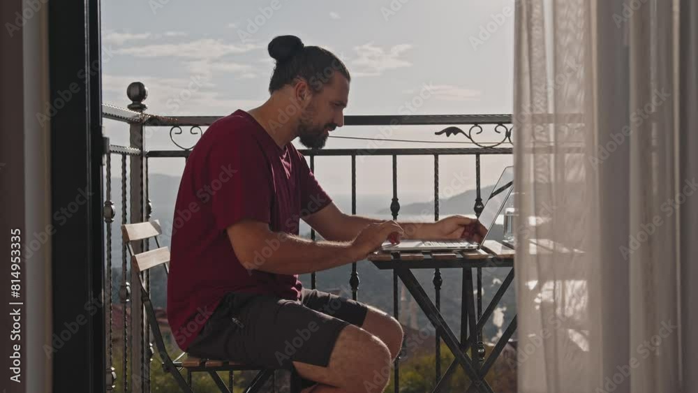 A man works on a computer while on a balcony in a tourist town on the sea A man works on a computer while on a balcony in a tourist town on the sea