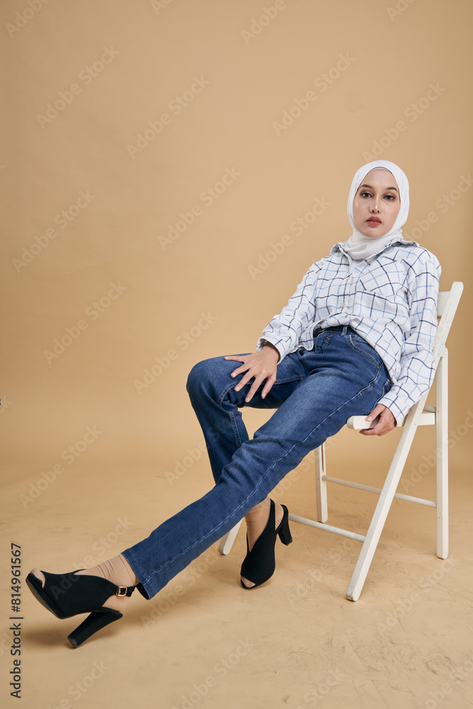 Fototapeta premium Portrait of an Asian model in white shirt and blue jeans with hijab sitting on a white chair isolated over beige background. Stylish Muslim female hijab fashion lifestyle concept.
