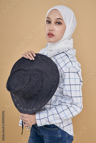 Portrait of an Asian hijab model in white shirt and blue jeans wearing cowboy hat isolated over beige background. Stylish Muslim female hijab fashion lifestyle concept. photo