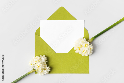 Green envelope with clean white sheet of paper for your text and spring flowers white daffodils on gray background. Invitation to the holidays. Flat lay.