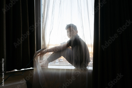 Boy sitting in the window, hiding behind a sheer curtain.  photo