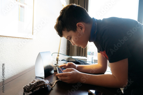Boy leaned over a desk playing a game on his phone.  photo