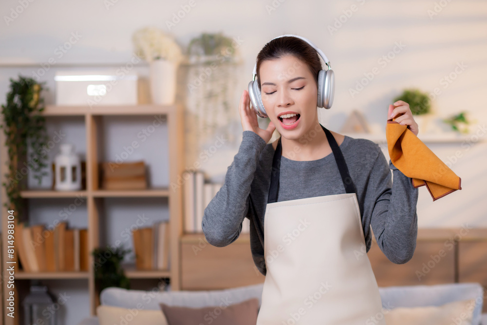 Young asian woman hold fabric while listening to music with headphones in living room, maid doing housework enjoying music and sing while dusting at home, lifestyle concept.