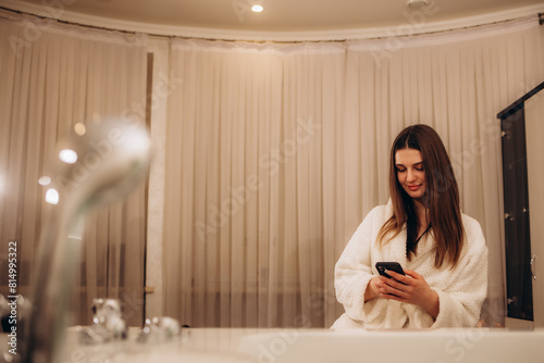Smiling young indian woman in white bathrobe using smartphone after shower, sitting on bathtub in bathroom, brushing teeth, holding phone, checking social network, reading message, good news