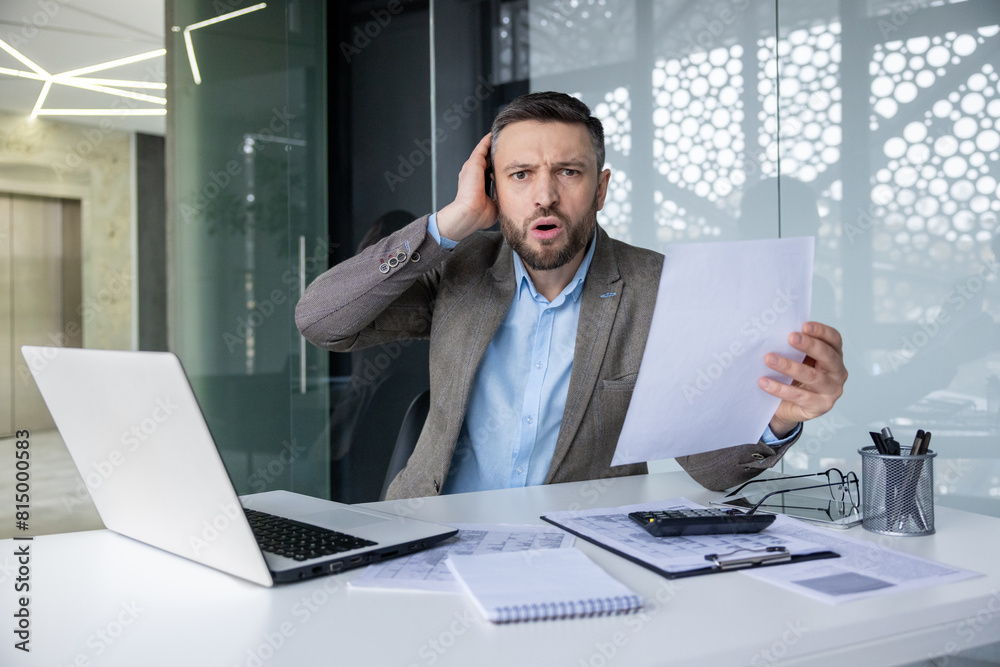 Shocked businessman holding a document while sitting at his desk in an office. He appears surprised and worried, surrounded by paperwork and a laptop. Shocked businessman holding a document while sitting at his desk in an office. He appears surprised and worried, surrounded by paperwork and a laptop.