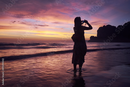 Back view of woman standing on beach taking camera photo of the vibrant sunset colours and landscape at Railey Beach, Thailand