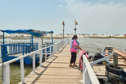 A senior African American woman with walking stick and wearing life jacket standing on a pier looking towards a sandy beach with blue sky and canoe in the background, Ghana