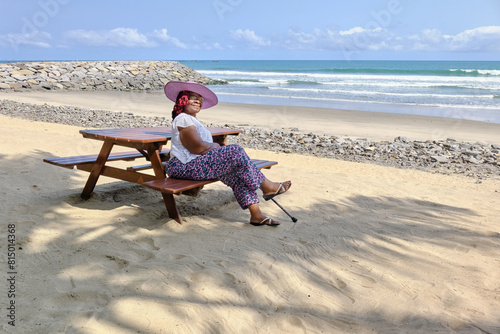 Senior African American woman wearing a purple hat sitting at a table and bench on the golden sandy Anomabo beach smiling and looking at the camera with the blue ocean in the background, Ghana