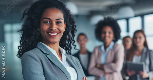 Professional young businesswoman smiles confidently at the camera with her diverse team blurred in the background, symbolizing leadership, teamwork, and workplace empowerment