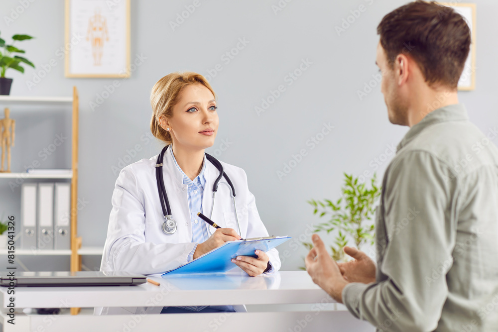 Young man patient talking to female blonde friendly doctor in her office in clinic during medical examination. Woman physician listening to the patient's complaints. Healthcare and medicine concept. Young man patient talking to female blonde friendly doctor in her office in clinic during medical examination. Woman physician listening to the patient's complaints. Healthcare and medicine concept.