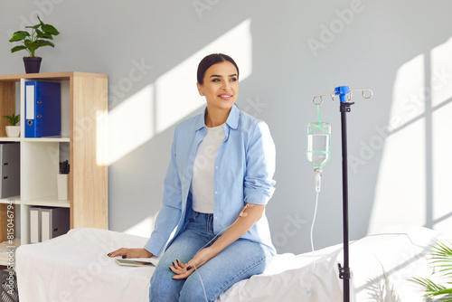 Foto Young happy smiling woman sitting on the couch in medical clinic looking at the window while receiving IV drip infusion and vitamin injection therapy in her blood