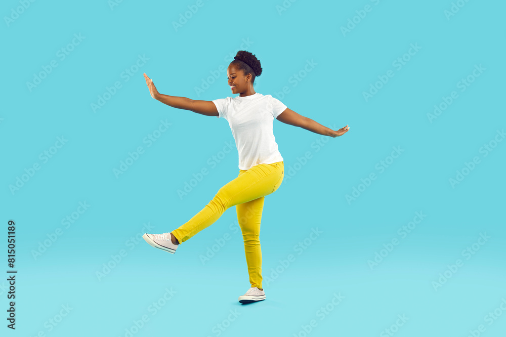 Full body shot of happy funny young African American woman wearing white T shirt and yellow pants pretending to be ballerina and dancing isolated on blue color background Full body shot of happy funny young African American woman wearing white T shirt and yellow pants pretending to be ballerina and dancing isolated on blue color background