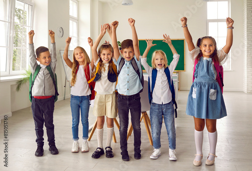 Childhood and school. Portrait of funny group of classmates from elementary school having fun in classroom. Smiling boys and girls with backpacks on their shoulders stand in row with raised hands.
