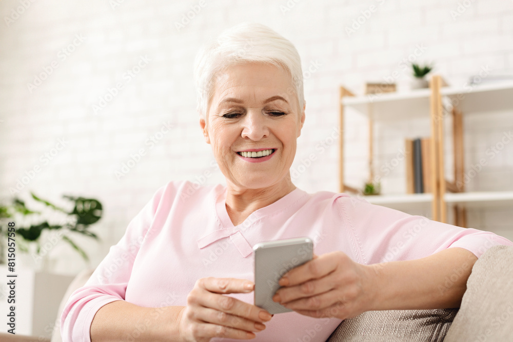 Senior woman is sitting on a couch, holding a cell phone and looking at the screen. She appears focused on the phones display, with her legs crossed and a relaxed posture. Senior woman is sitting on a couch, holding a cell phone and looking at the screen. She appears focused on the phones display, with her legs crossed and a relaxed posture.