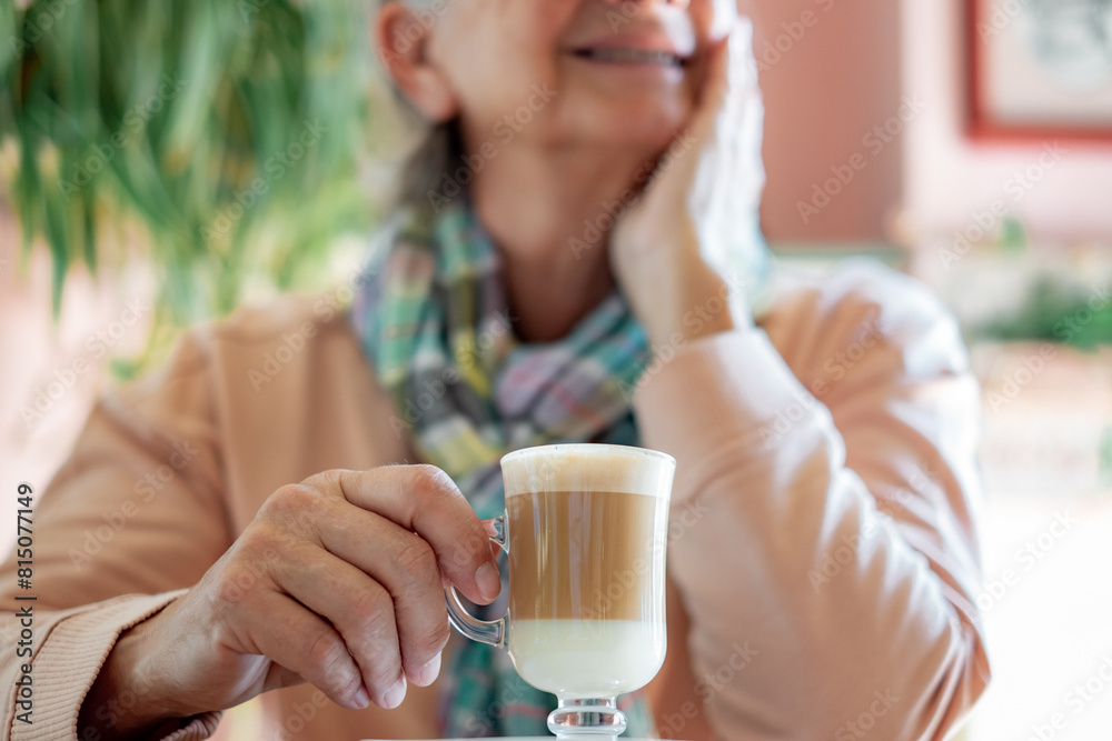 Senior woman sitting at cafe table holding a glass cup filled with coffee and milk. Elderly lady enjoying relaxed break with hot drink Senior woman sitting at cafe table holding a glass cup filled with coffee and milk. Elderly lady enjoying relaxed break with hot drink