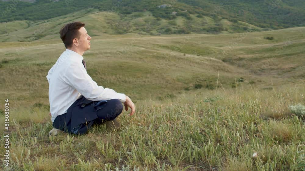 Adult young man in business suit in profile among the hills in relaxed pose and looking at sunset. The man has taken off his jacket and is sitting on the grass. Looks at dawn sun rise up. Adult young man in business suit in profile among the hills in relaxed pose and looking at sunset. The man has taken off his jacket and is sitting on the grass. Looks at dawn sun rise up.
