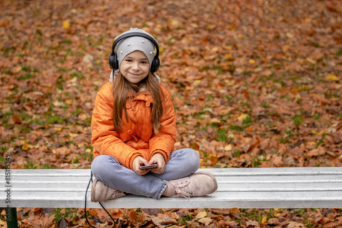 A girl listens to music on headphones in an autumn park while sitting on a white bench.