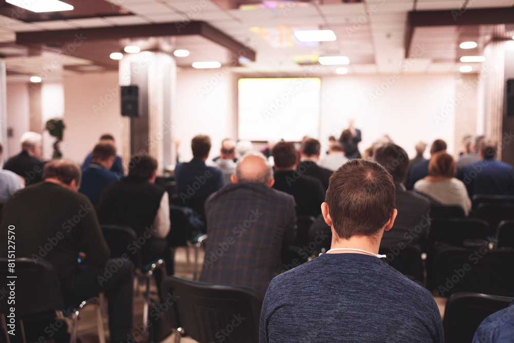 A large group of people are sitting in a room, listening to a speaker. Scene is serious and focused, as everyone is attentive to the speaker A large group of people are sitting in a room, listening to a speaker. Scene is serious and focused, as everyone is attentive to the speaker