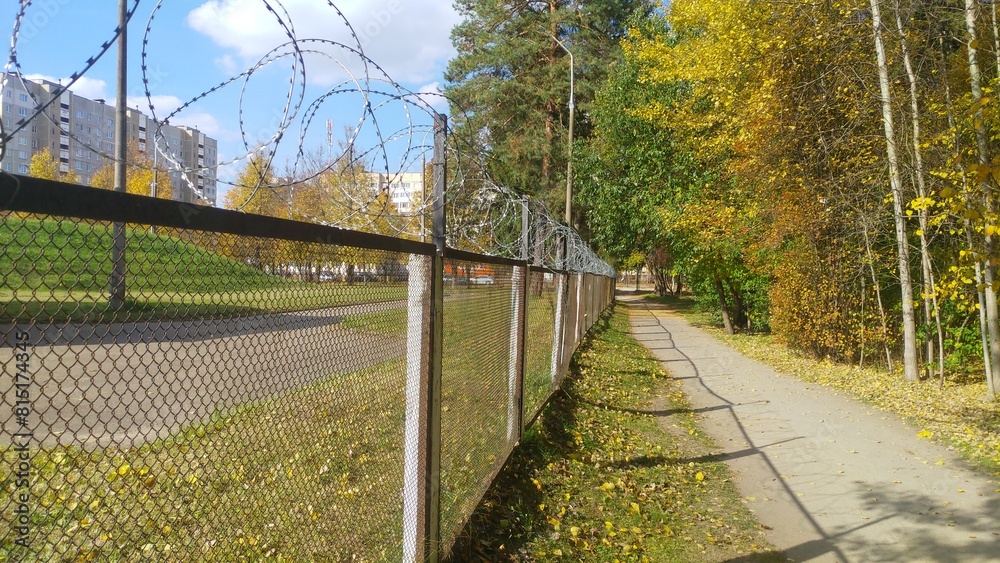 On one side of the unpaved walkway are the trees of a city park. On the other side there is a metal mesh fence with concrete posts and barbed wire stretched over the top. Sunny autumn weather On one side of the unpaved walkway are the trees of a city park. On the other side there is a metal mesh fence with concrete posts and barbed wire stretched over the top. Sunny autumn weather