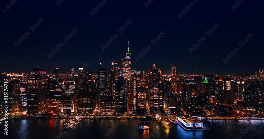 Flying closer to a stunning skyline of New York at the waterfront of the East River at night. Lights of the planes flashing in the blue sky above the cityscape.