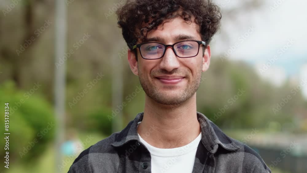 Portrait of a young handsome man in glasses close up, standing in an outdoor city park, looking at the camera and smiling with a calm, confident face