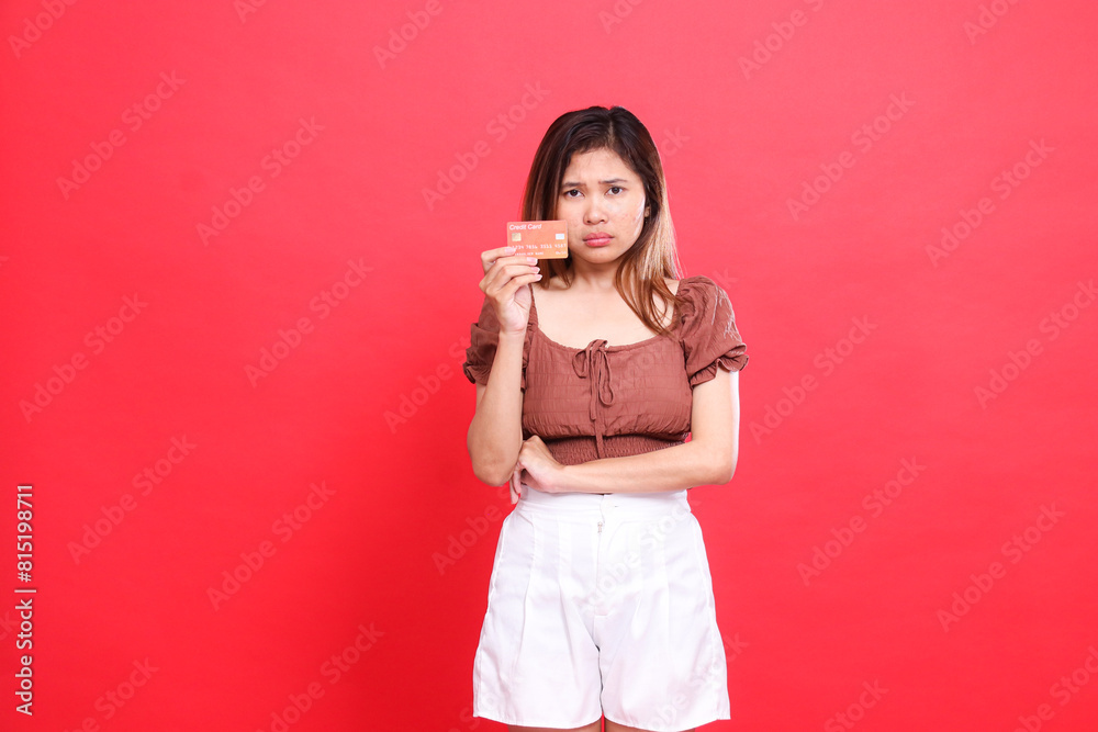 sad indonesia woman holding her arms from the side of a debit credit card wearing a brown blouse on a red background. for transaction, technology and advertising concepts sad indonesia woman holding her arms from the side of a debit credit card wearing a brown blouse on a red background. for transaction, technology and advertising concepts