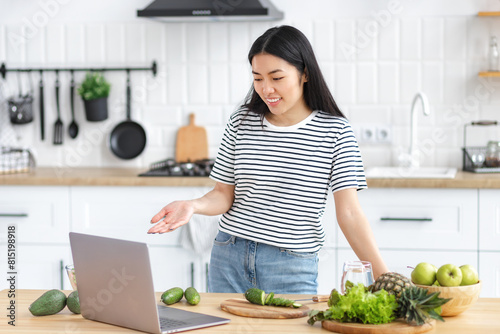 Young woman cooking healthy food and communicates online using laptop standing at home in the kitchen