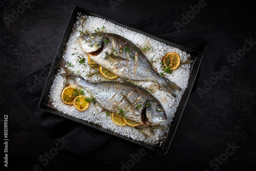 Two fresh steamed gilthead seabreams in salt crust with lemon slices and herbs served as top view in an old metal sheet © HLPhoto